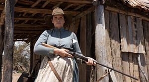Women standing on a porch of a wooden house in clothing from 1800s holding a gun for the new film The Drovers Wife: The Legend Of Molly Johnson